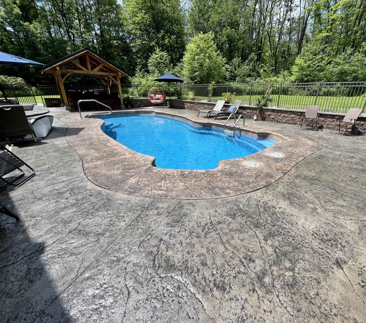 A stamped concrete pool deck wrapping an in-ground pool on a wooded lot